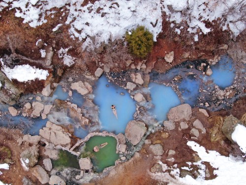 Aerial view of natural hot springs surrounded by rocks, snow, and trees, with two people relaxing in the water.