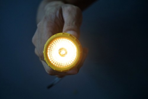 A hand holding a yellow flashlight with a bright light shining from the front against a dark background.