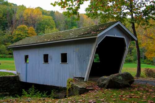 A weathered blue covered bridge surrounded by autumn foliage and grassy fields.