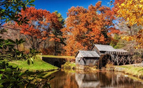 A scenic view of a wooden mill by a pond, surrounded by vibrant autumn foliage and a clear blue sky.