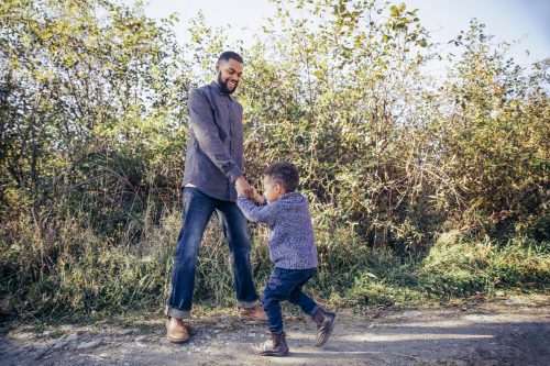 A man and a young boy joyfully dance together outdoors, surrounded by greenery and sunlight.