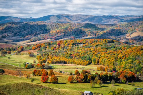 A scenic view of rolling hills and valleys adorned with vibrant autumn foliage under a partly cloudy sky.