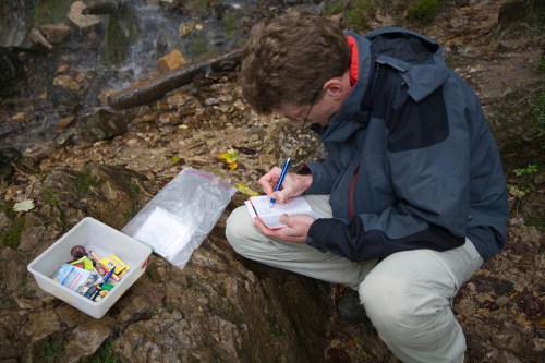 A person crouches by a stream, writing notes while surrounded by a container of supplies on the ground.