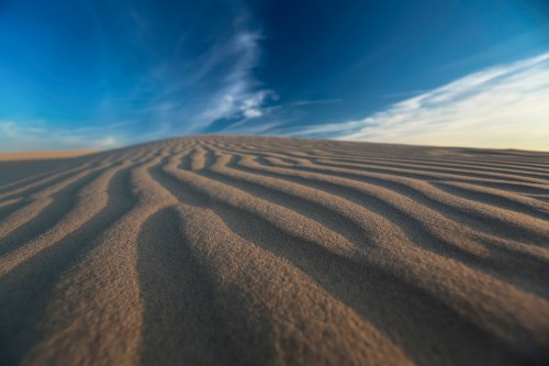 Close-up of rippled sand dunes under a blue sky with wispy clouds, creating a serene desert landscape.