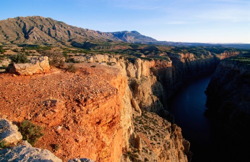 A stunning canyon landscape with steep cliffs, a winding river, and mountains in the background under a clear blue sky.