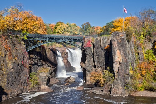 A scenic view of a waterfall with a bridge, surrounded by colorful autumn foliage and a clear blue sky.
