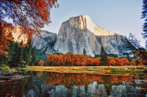 A stunning view of a mountain reflecting in a calm lake, surrounded by vibrant autumn foliage and clear blue skies.