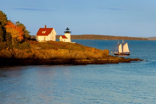 A scenic coastal view featuring a lighthouse, a white house, and a sailboat near rocky shores under a clear sky.