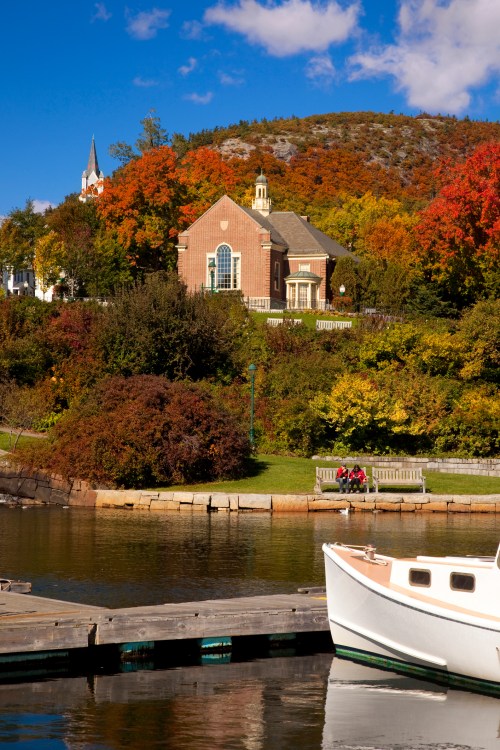 A serene waterfront scene featuring a boat, colorful autumn foliage, and a charming building on a hillside.