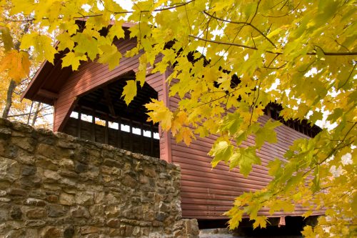 A wooden covered bridge surrounded by vibrant autumn leaves and a stone wall.