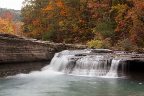 A serene waterfall cascades over rocky ledges, surrounded by vibrant autumn foliage and calm waters.
