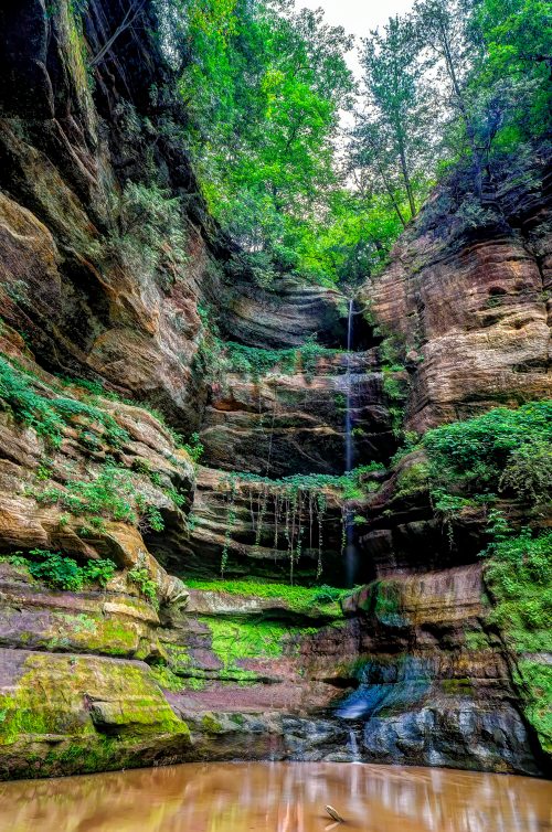 A serene waterfall cascades down rocky cliffs, surrounded by lush greenery and reflected in a calm pool below.