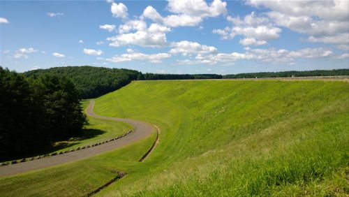A winding road curves along a grassy hillside under a blue sky with fluffy clouds. Trees line the background.