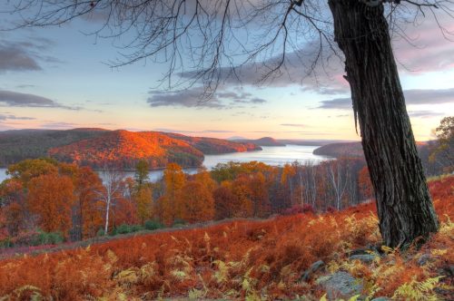 A scenic view of a lake surrounded by vibrant autumn foliage and rolling hills under a colorful sky.