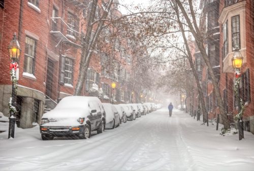 A snowy street lined with brick buildings and parked cars, with a lone figure walking in the distance amidst falling snow.