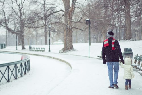 A man and a child walk hand in hand along a snowy path, surrounded by trees and benches in a winter park.