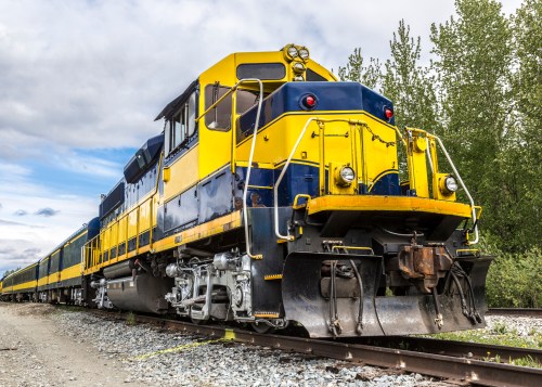 A bright yellow and blue train engine parked on tracks, surrounded by greenery under a cloudy sky.
