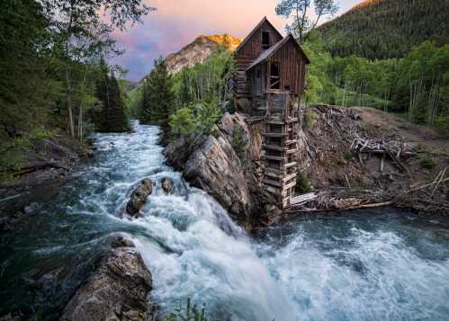 A rustic wooden cabin perched on a rocky outcrop beside a rushing river, surrounded by lush greenery and mountains.