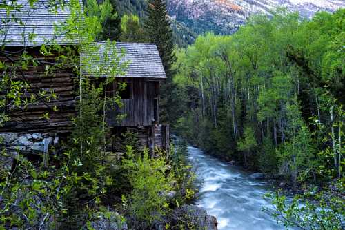 A serene river flows beside a rustic wooden cabin, surrounded by lush green trees and mountains in the background.