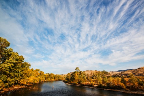 A serene river flows through a landscape of autumn trees under a blue sky with wispy clouds.