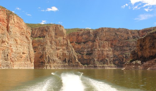 A scenic view of towering cliffs beside a calm lake, with a clear blue sky and white boat wake in the foreground.