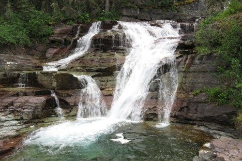 A cascading waterfall flows over rocky cliffs into a serene pool, surrounded by lush greenery.