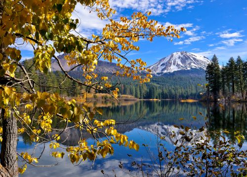 A serene lake reflects autumn foliage and a snow-capped mountain under a clear blue sky.