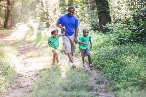 A man walks hand-in-hand with two children along a sunny, wooded path, surrounded by greenery.