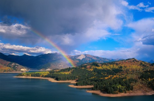 A vibrant rainbow arcs over a serene lake surrounded by colorful autumn foliage and mountains under a partly cloudy sky.