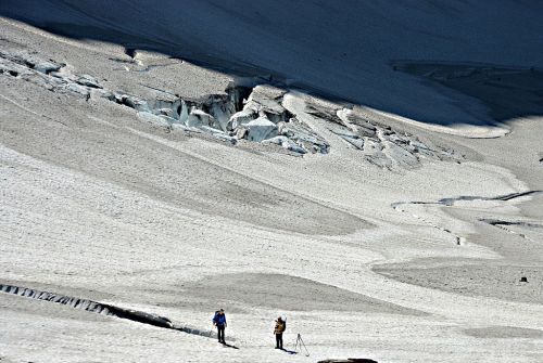 Two hikers explore a vast, snowy landscape with glacial formations under a clear blue sky.