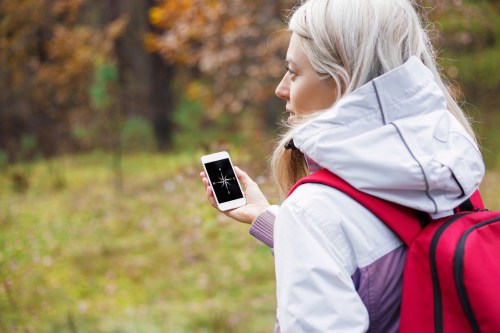 A woman with a backpack checks her phone while walking in a forested area during autumn.
