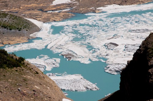Aerial view of a turquoise lake with ice and snow patches surrounded by rocky terrain and sparse vegetation.