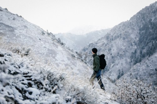 A person in winter attire stands on a snowy hillside, gazing at distant mountains in a serene landscape.