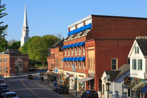 A quaint town scene featuring brick buildings, shops, and a church steeple in the background under a clear blue sky.