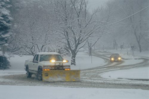 A snowy scene with a pickup truck equipped with a plow clearing a road while another car navigates the snowy curve.