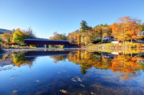 A serene autumn scene featuring a covered bridge reflected in a calm river, surrounded by colorful fall foliage.