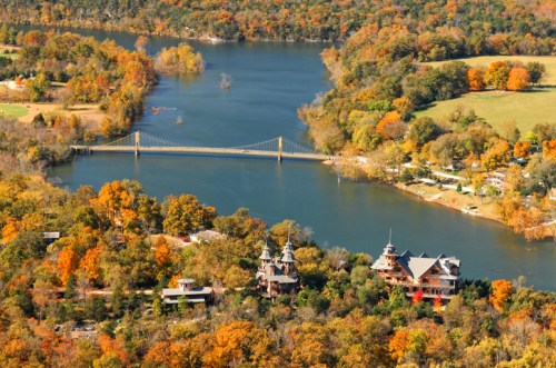 Aerial view of a river surrounded by autumn foliage, featuring a bridge and two distinctive houses along the shore.