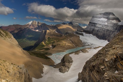 A breathtaking mountain landscape featuring rugged peaks, a glacier, and a turquoise lake under a partly cloudy sky.