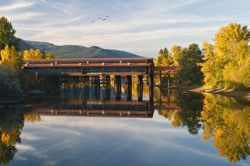 A serene river scene with a modern building reflecting on the water, surrounded by autumn trees and mountains.