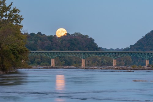 A full moon rises over a green bridge spanning a calm river, surrounded by trees and hills at dusk.