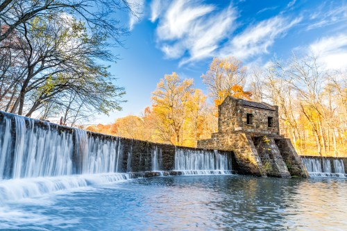 A scenic view of a stone structure beside a waterfall, surrounded by autumn trees and a clear blue sky.