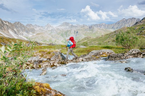 A hiker with a red backpack crosses a rocky stream in a mountainous landscape under a blue sky.