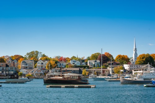 A scenic harbor with boats, colorful autumn trees, and a church steeple against a clear blue sky.