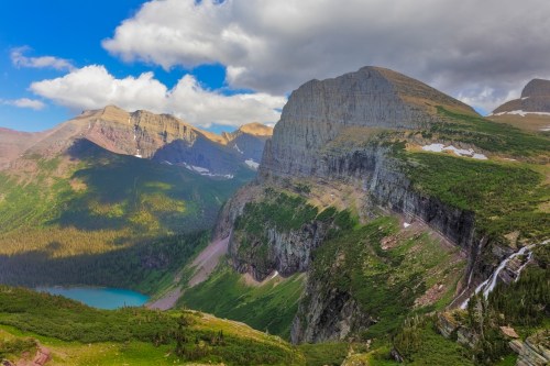 A stunning mountain landscape with lush greenery, a lake, and dramatic cliffs under a partly cloudy sky.