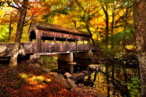 A wooden covered bridge over a serene stream, surrounded by vibrant autumn foliage and colorful trees.