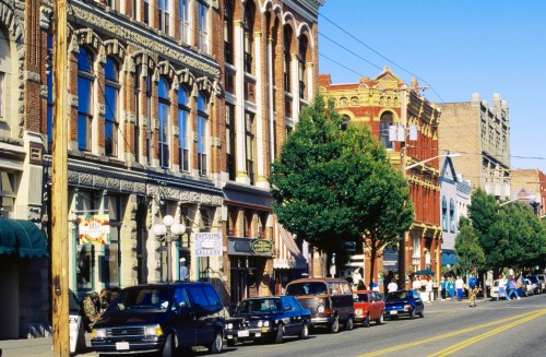 Historic buildings line a sunny street, with parked cars and pedestrians enjoying the vibrant atmosphere.
