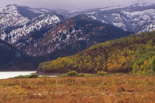 A scenic view of mountains with snow, colorful autumn trees, and a calm lake in the foreground.