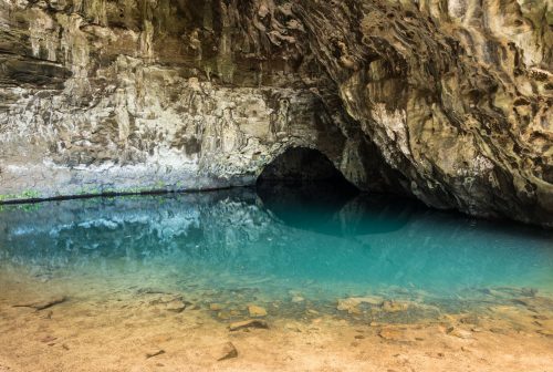 A serene cave entrance with turquoise water reflecting the rocky walls, surrounded by earthy tones and greenery.