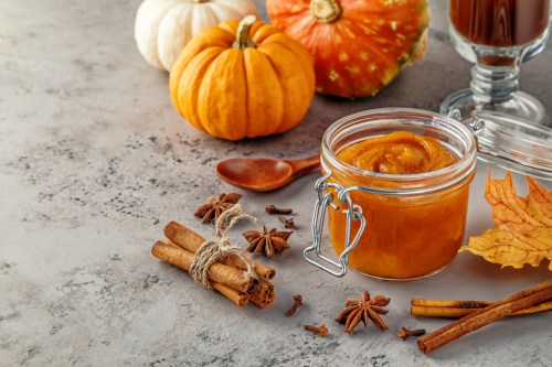 A jar of pumpkin puree surrounded by pumpkins, cinnamon sticks, and autumn leaves on a textured surface.