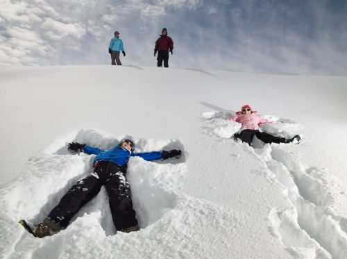 Two people making snow angels in a snowy landscape, while two others stand on a hill in the background.